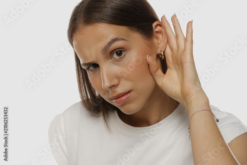 Portrait of a young woman listening carefully with hand near her ear, natural beauty, no makeup, on white background.