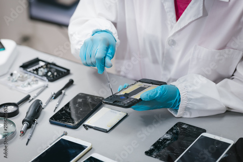 Technician in protective gloves repairs a damaged smartphone on a workbench with tools and broken phone screens.