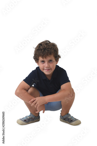 front view of a 6-year-old boy dressed in summer clothes, squatting and looking at camera on white background.