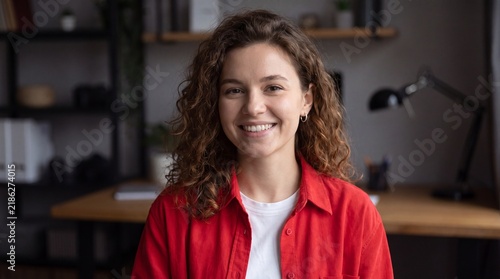 Portrait of a young woman with curly brown hair wearing a red shirt in a home office setting