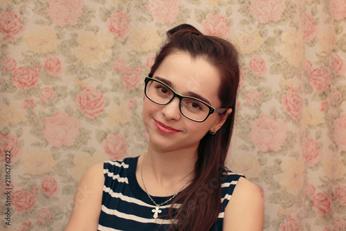 A young woman with glasses and a striped top smiling at the camera against a floral background. Portrait, casual style, friendly expression. cross around the neck. High quality photo