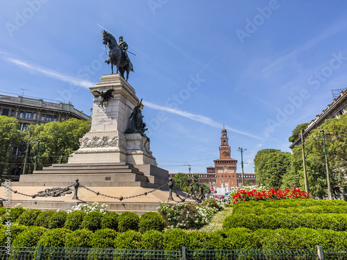 Fotografía Giuseppe Garibaldi's equestrian statue stands in Milan's Piazza Cairoli, with Castello Sforzesco rising beyond the gardens under a bright blue sky