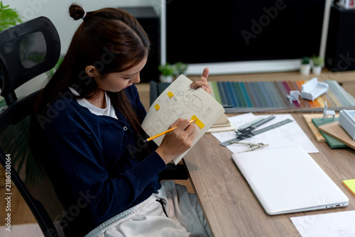 Asian Female Furniture Designer Reviewing Technical Sketches and Color Samples at Contemporary Home Office Desk