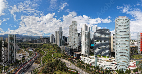 Aerial panoramic view of the Santa Fe skyline and Parque La Mexicana in Mexico City under a blue cloudy sky.