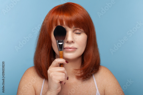 Принти на полотні Calm mature redhead woman holding large black makeup brush in front of her face