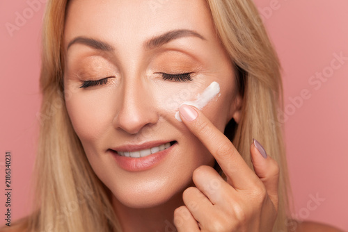 Mature blonde woman smiling gently with closed eyes while applying anti-aging moisturizer with her finger on cheek isolated on pink background