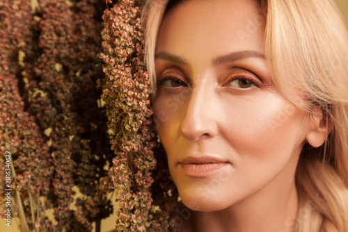 Intense gaze of blonde woman behind autumn vegetation looking straight at camera and showing perfect skin isolated over yellow background