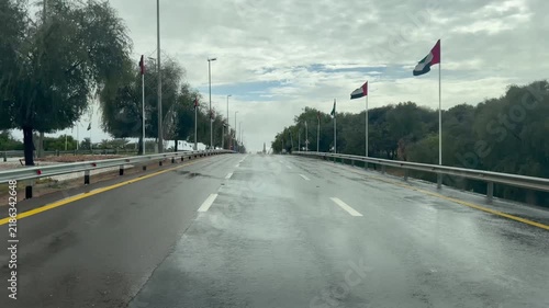A rain‑soaked multi‑lane UAE highway lined with national flags at half‑mast beneath a grey, overcast sky.