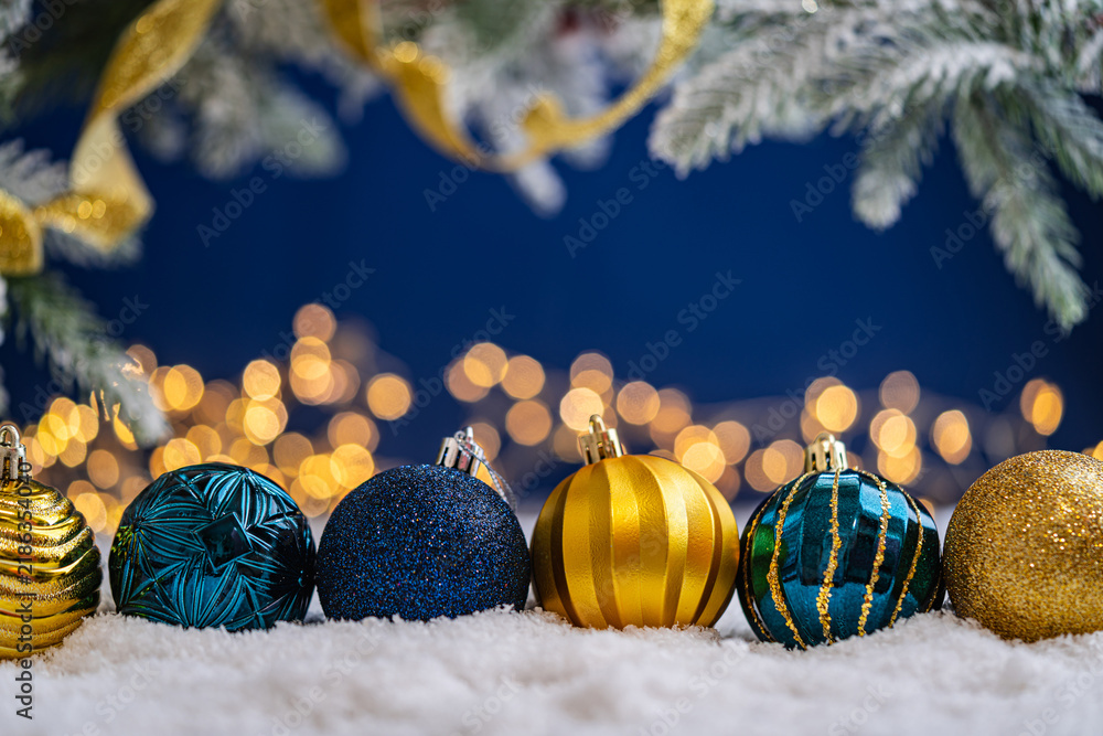 Naklejka premium A Christmas background featuring a row of teal and navy baubles with etched patterns and gold accents on snow, framed by frosted pine and gold ribbon, set against deep blue with lights.