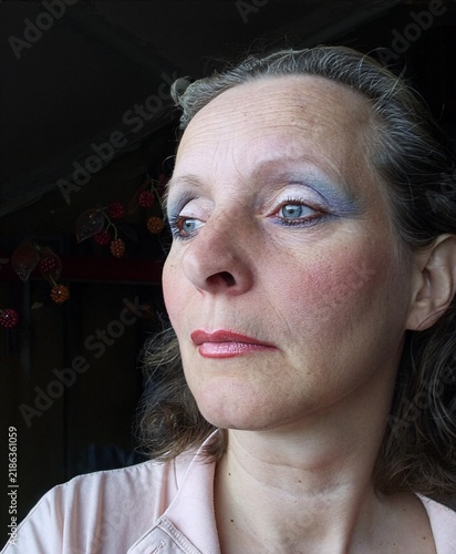 Woman of 50 with makeup applied, including blue eyeshadow, pink blush, and pink lipstick. Her hair is loose and she wears a light-colored top. 