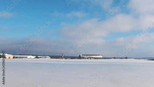 A wide view captures a snow-covered field stretching across the landscape. A barn stands in the distance, surrounded by fresh snow. The sky is blue with some clouds, creating a winter scene. 
