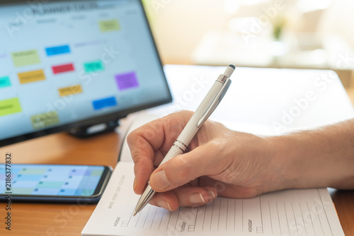 Person's hand writing daily tasks and appointments in a paper planner, cross-referencing with a digital calendar displayed on a smartphone and tablet for efficient time management and organization
