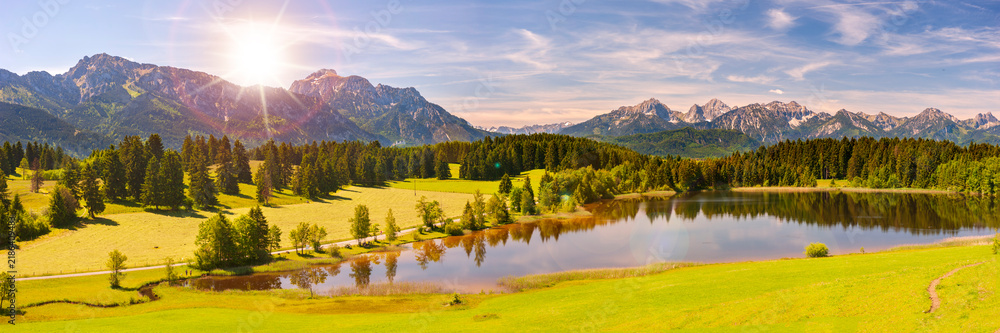 Obraz premium Panorama Landschaft im Allgäu mit saftigen Frühlingswiesen und Blick über den Hegratsrieder See zu den Alpen - FOTOMONTAGE