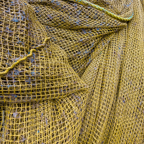 A detailed close-up of piled, textured yellowish-brown commercial fishing nets on a deck, featuring complex mesh patterns, knots, and a visible yellow hardware ring.