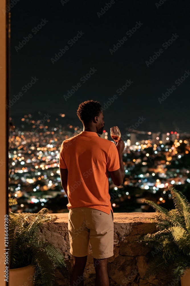 Obraz premium Rear view of man with wine glass on balcony overlooking illuminated cityscape at night for lifestyle banner and romantic evening background