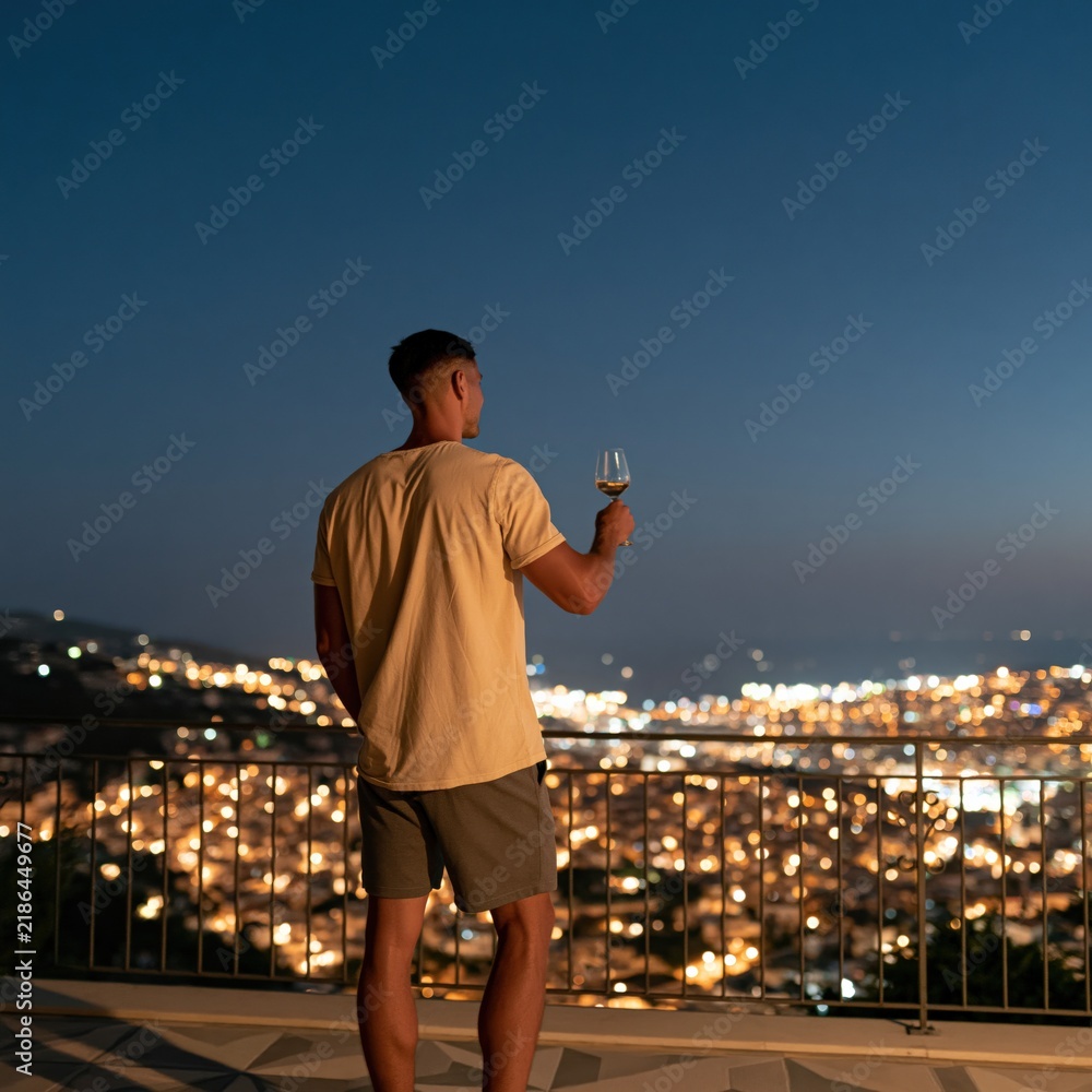 Obraz premium Man on balcony at night overlooking city lights holding a wine glass for rooftop background and travel lifestyle banner design