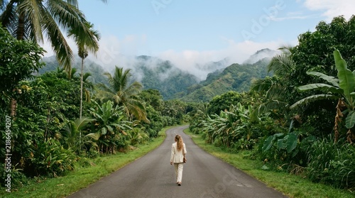 Woman in a light suit walking along a tropical road, lush jungle and misty mountains in the background.
