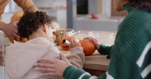 African American family carving pumpkins in kitchen; dad bringing pumpkin, child tracing with tool