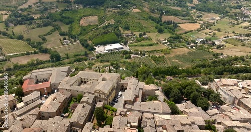 High aerial view captures the edge of a historic Italian hilltop town overlooking a lush green valley, showcasing traditional stone architecture, tile roofs, and scenic rolling farmland.