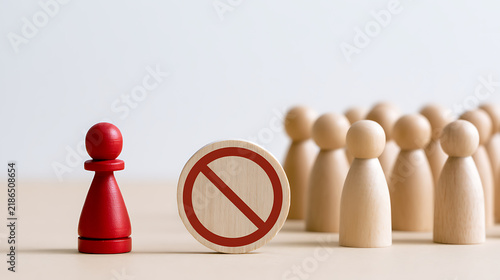 Red pawn standing separate from a group of wooden figures behind a prohibition sign depicting social exclusion, discrimination, and rule