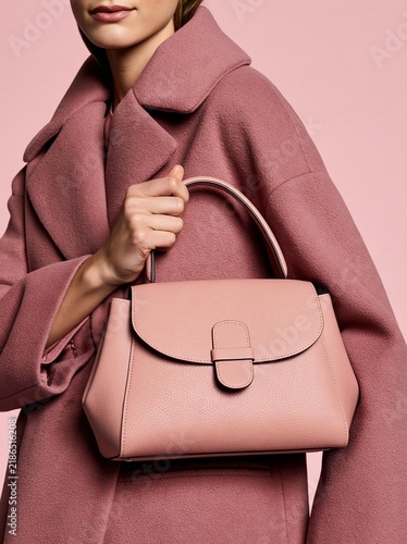 Woman holding a pink handbag in her right hand. she is wearing a long, pink coat with a high collar and long sleeves. the coat is made of a soft, textured fabric and has a button closure on the front.