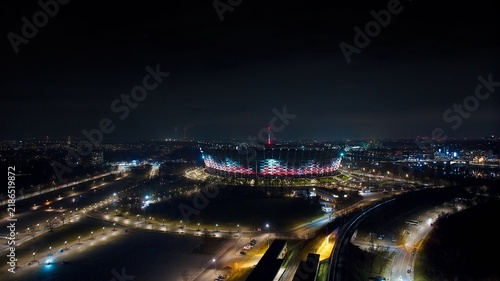 Aerial drone capture of Warsaw at night with the glowing PGE Narodowy, Świętokrzyski Bridge, and bustling streets.