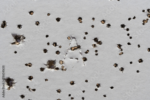 Macro photograph of a textured white shooting target paper densely perforated by multiple shotgun pellets featuring a distinct central letter shape.