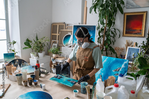 Woman wear respirator and protective gloves applies colored epoxy resin to abstract panel in home loft workshop, creating unique handmade artwork to monetize artistic hobby through small business.