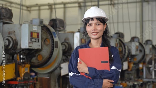 Female engineer wearing hard hat and protective uniform holding red folder inside manufacturing factory, confident industrial worker among heavy machinery, metalworking equipment, and industrial