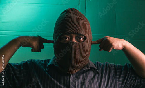 Stubborn Indonesian adult male wearing a brown balaclava covering the head while plugging both ears with index fingers to block loud noise. The anonymous Asian man dressed in a dark