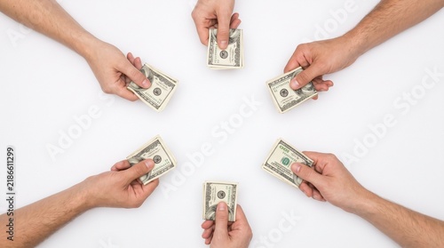Top view of six hands arranged in a circle on a plain white background, each holding a stack of folded 100 US dollar bills facing inward.