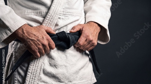 Close up of a martial artist's hands tying a black belt around their traditional uniform against a dark background