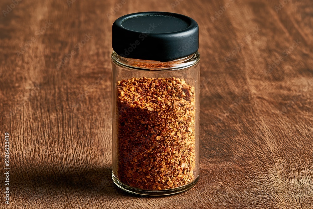 Fototapeta premium Chili pepper flakes and dry seasoning blend in shaker bottle stands on wooden table for restaurant food flavoring needs.