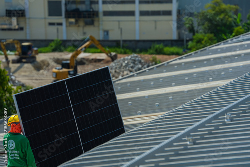 Konstfotografi A solar installation engineer in a white helmet and high visibility vest precisely carries a single solar panel across the roof past an excavator below high on an industrial warehouse roof