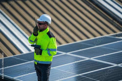 Obraz na płótnie A solar installation engineer in a white helmet and high visibility vest precisely walks the golden evening array with a radio in hand high on an industrial warehouse roof in a Thai city
