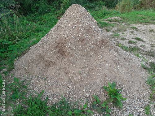 Pile of Gravel and Sand on Ground. A sand-and-gravel mix for construction or landscaping. A mound of stone and sand material lies next to patches of green grass.
