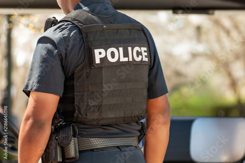 A male police officer wears a taser and a handgun. The law enforcement officer wears a black bulletproof vest, a black shirt, and dark pants. The word police is on his back. 