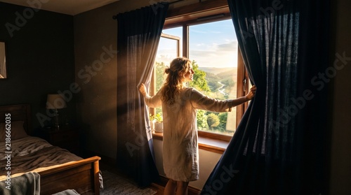 Woman opening curtains in bedroom to reveal a beautiful sunlit landscape with rolling hills and a river