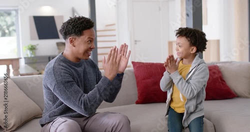 African American man raising palms, boy mirroring clapping and high-fives on sofa for bonding