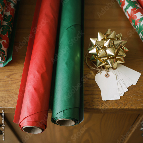 Red and green wrapping paper rolls with a gold star ornament and blank gift tags on wood