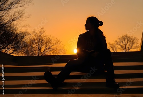 A silhouette of a student sitting on library steps at sunset, warm dramatic backlight