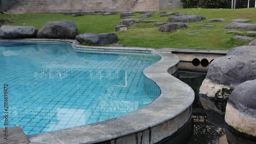Natural pool curves beside large rocks. Clear water reflects sky and surrounding greenery. Stone border blends with landscaped garden setting. Ideal for vacation, spa, or wellness imagery