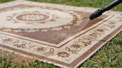 Rug cleaning process involves spray nozzle washing patterned textile brown cream floor covering outdoors on green grass lawn.