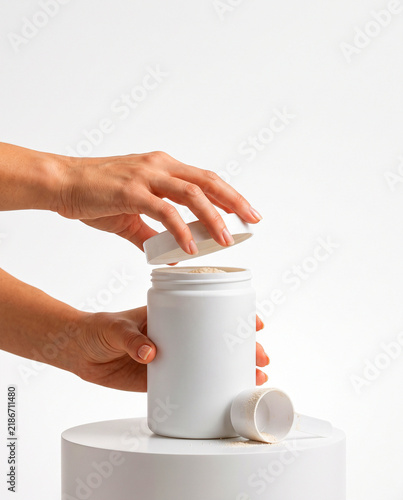 Hands Opening White Supplement Powder Jar with Scoop on Podium
