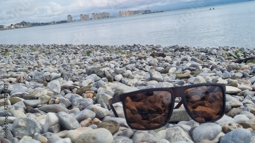 Sunglasses resting on a pebble beach with the ocean in the background. The sunglasses have dark lenses and a brown frame, reflecting the cloudy sky and buildings in the distance
