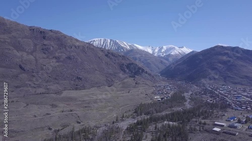 Mountain settlement occupies valley floor below snow covered mountain range. Aerial landscape passes over clustered rooftops, winding terrain, and dark tree stands beneath cloudless blue sky.