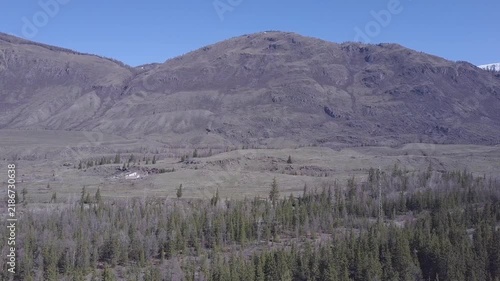 Wilderness mountain range overlooks dense evergreen forest and wide valley floor. Weathered ridges and clear sky define remote landscape while aerial movement follows natural terrain.