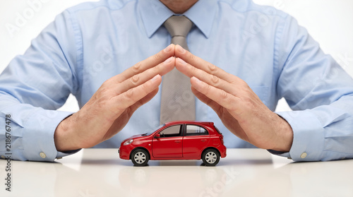 Professional businessman in light blue shirt holding hands in a protective gesture over a small red toy car model, symbolizing vehicle insurance, safety, and security concept on white background