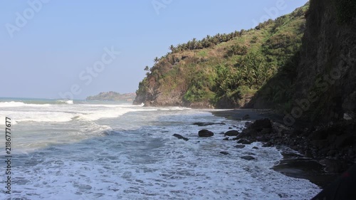 Rugged cliffs meet crashing waves at a wild, rocky shore. Green foliage climbs steep, weathered rock faces beside the sea. Foamy surf crashes against jagged stones with force and rhythm