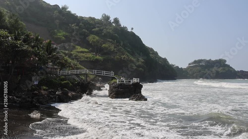 Waves crash against rocky shore under bright sky. Green cliffs rise behind a weathered wooden walkway. Palm trees dot the rugged coastline. White foam dances as sea meets land with force